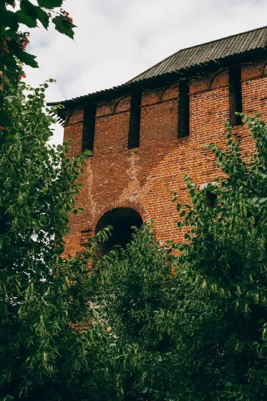 Краснокирпичная крепостная башня за зелеными ветвями деревьев.